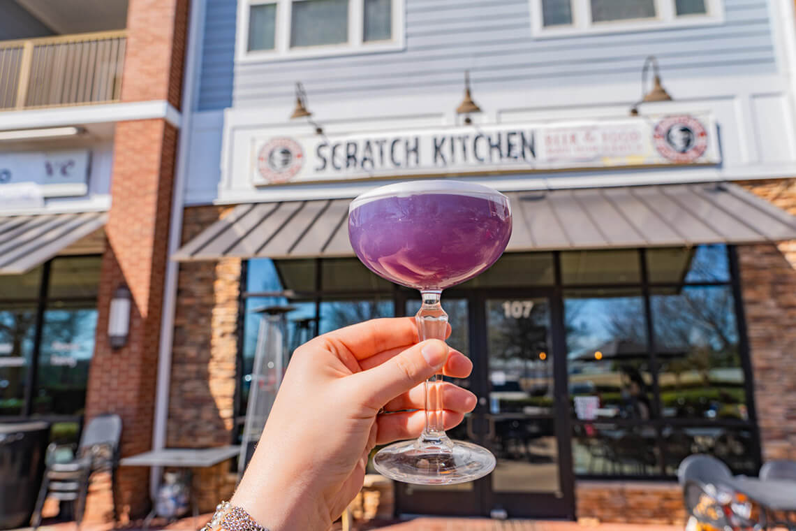 Hand holding a purple cocktail outside of Scratch Kitchen